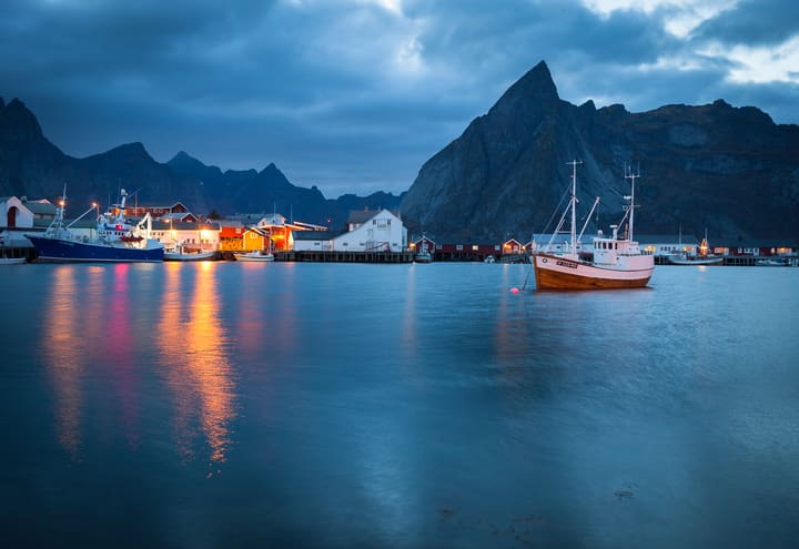 Fishing boats and lit cabins in Hamnøy harbour at dusk under dark Lofoten peaks